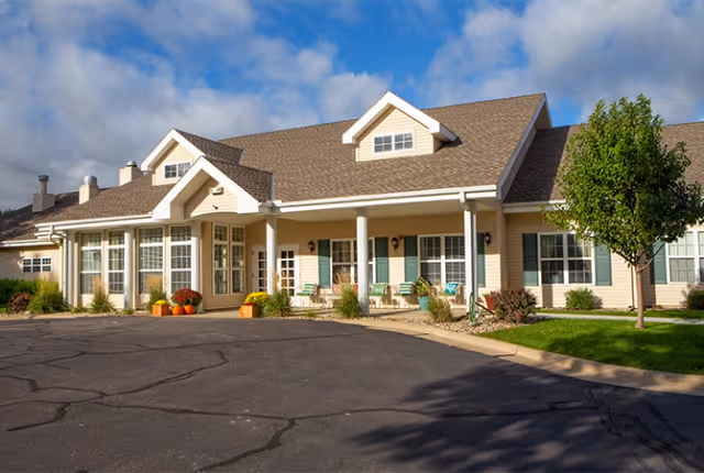 Single-story beige senior living building with a covered front porch, columns, seating and landscaping under a partly cloudy sky.