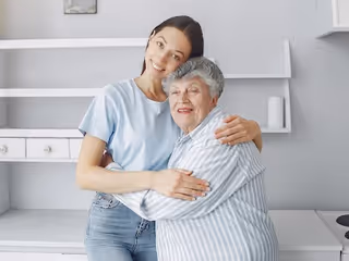 A young woman and an elderly woman hugging each other and smiling in a bright, modern kitchen setting.