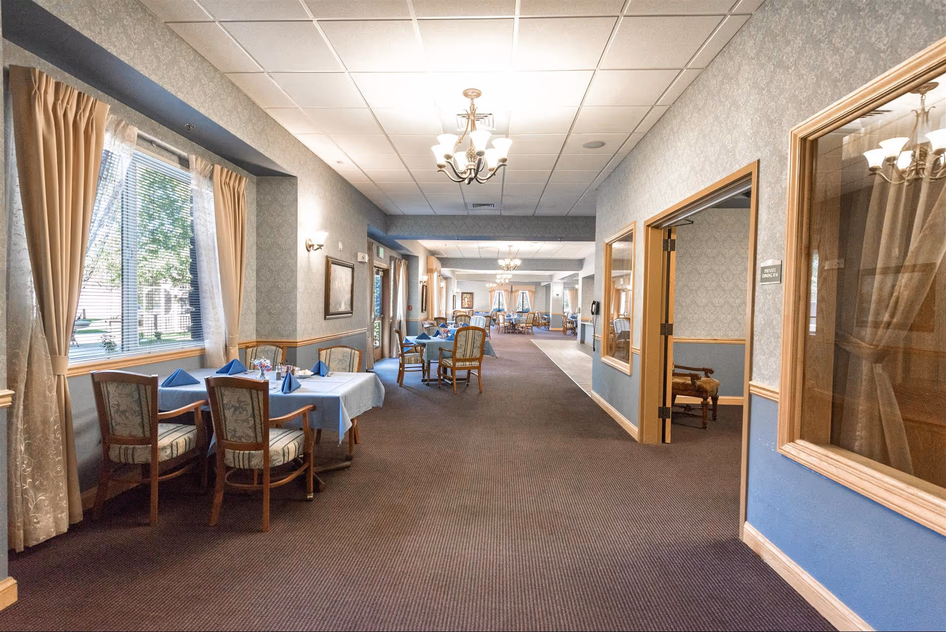Long carpeted dining corridor with tables set by windows, chairs, and chandeliers in a senior living facility.