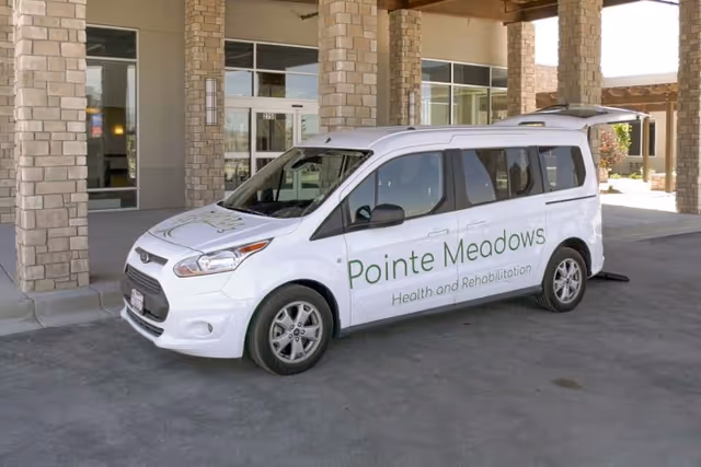 White van with the text 'Pointe Meadows Health and Rehabilitation' parked under a covered entrance of a building with stone pillars and large windows.