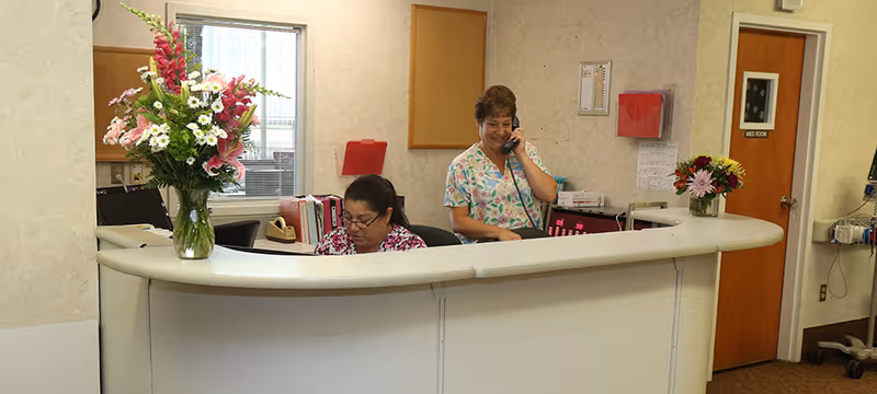 Reception area with two women behind a curved white desk. One woman is sitting and working on a computer while the other is standing and talking on the phone. There are two vases with colorful flowers on the desk, a window behind them, and a door labeled 'MED ROOM' to the right.
