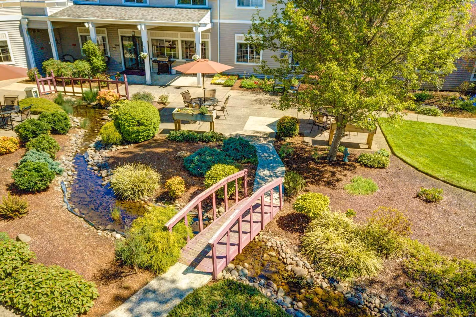 A landscaped outdoor garden area at The Oaks at Lebanon featuring a small wooden footbridge over a narrow stream with rocks, surrounded by various shrubs and plants. There are several patio tables with umbrellas and chairs on a concrete patio near the building entrance, which has a covered porch with white columns.