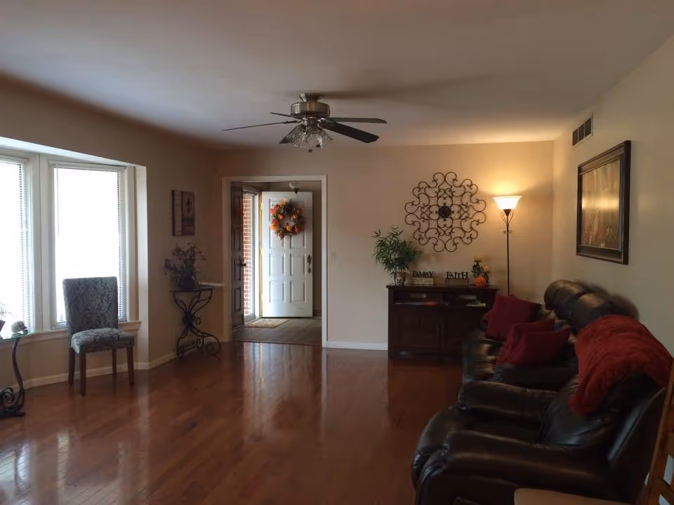 A living room with wooden flooring, a ceiling fan with lights, a large window with a patterned chair in front of it, a small table with a plant, and a dark leather sofa with red cushions and a red throw blanket. There is a wooden cabinet against the wall with decorative items and a floor lamp beside it. The entrance door is visible in the background with a fall-themed wreath hanging on it.