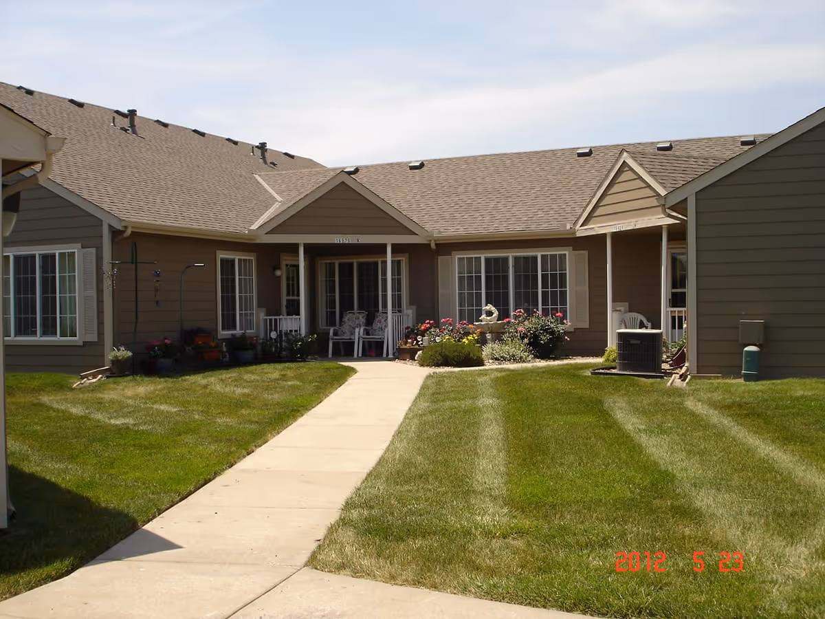 Exterior view of a single-story residential building with a beige exterior and multiple windows. A concrete walkway leads through a well-maintained lawn with striped mowing patterns to a small porch area with two chairs and potted plants. The sky is partly cloudy.