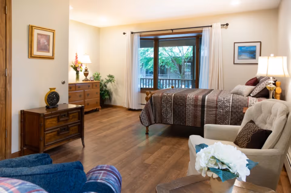 A cozy bedroom with a wooden bed covered in a patterned quilt, a beige armchair with a dark pillow, a wooden dresser with a lamp and flowers, a small wooden side table with a decorative vase, and a large window with white curtains showing green trees outside.