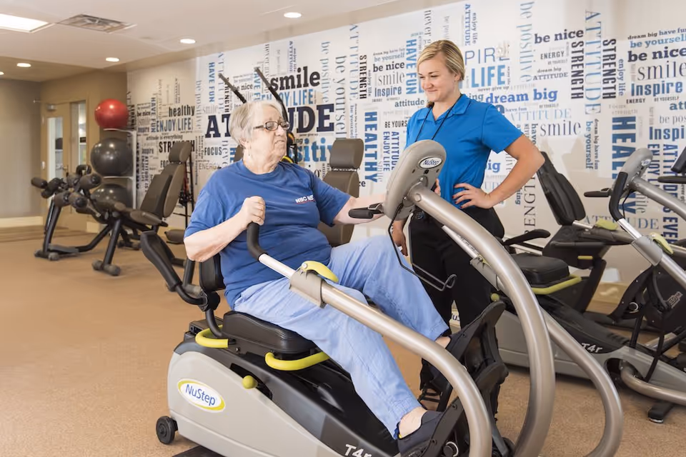 An elderly woman using a NuStep exercise machine in a fitness room, assisted by a staff member wearing a blue shirt. The room has exercise equipment and a wall decorated with motivational words like 'smile', 'attitude', and 'healthy'.