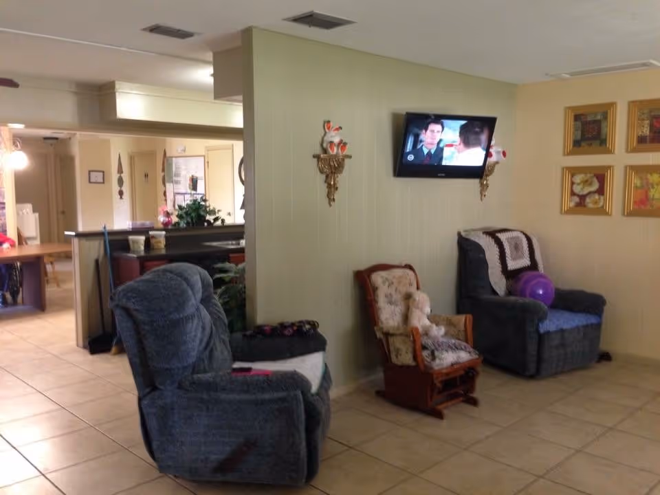 A cozy living room area with two upholstered recliner chairs and a small wooden rocking chair with a teddy bear on it. A flat-screen TV is mounted on a light green wall, showing a scene from a TV show or movie. The room has tiled flooring and some framed artwork on the wall. In the background, there is a partial view of a kitchen or dining area with a counter and some plants.