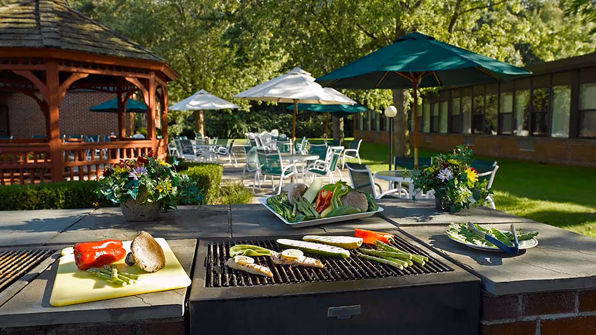 Outdoor barbecue grill with vegetables such as asparagus, bell peppers, and mushrooms cooking on it. In the background, there are tables and chairs with umbrellas set up on a patio area surrounded by greenery and a wooden gazebo.