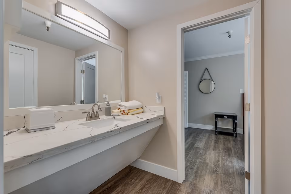Bathroom vanity with marble countertop, sink and large mirror, looking into an adjoining room with a round wall mirror and small table.