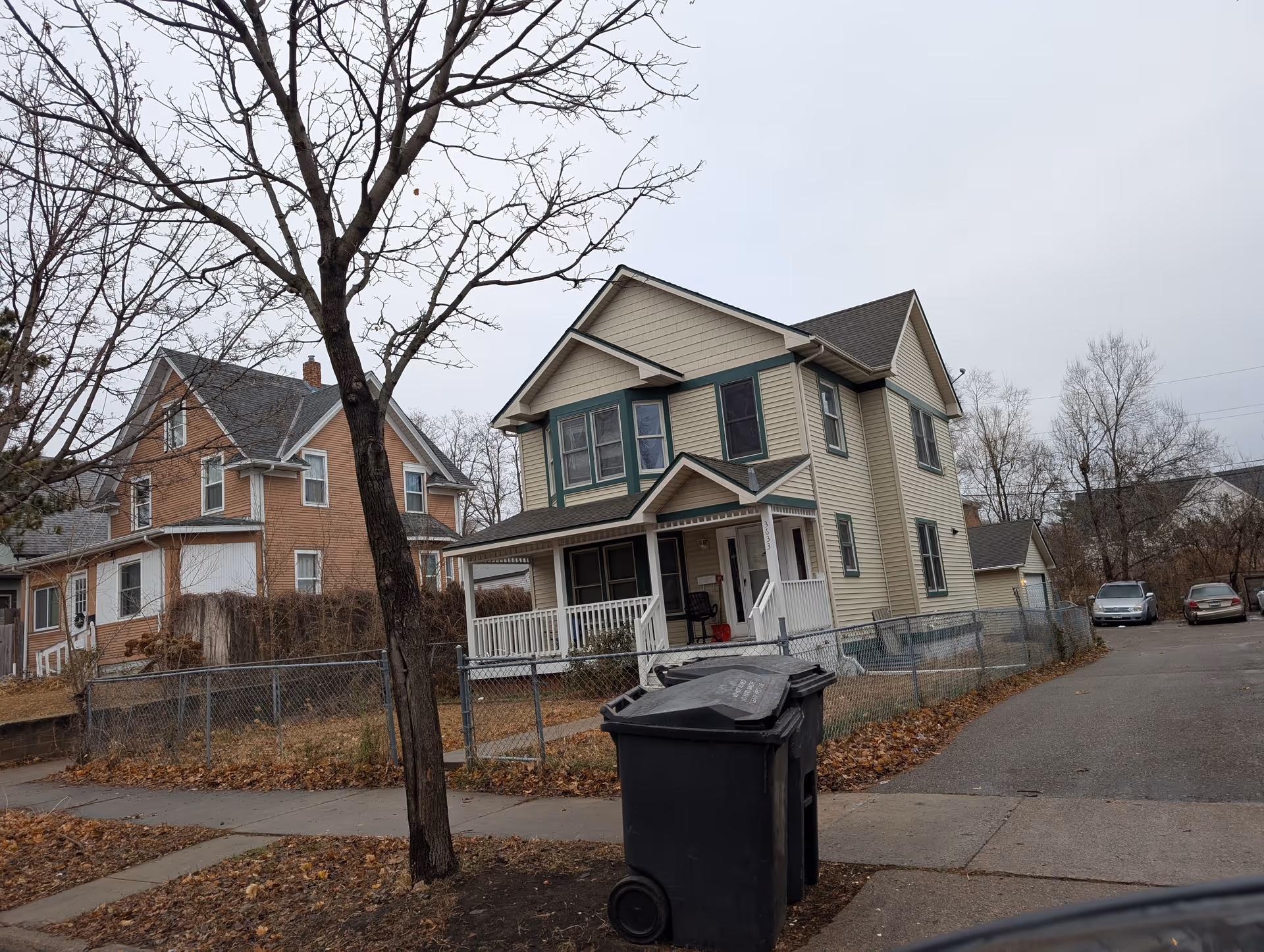 A two-story beige house with green trim and a front porch, situated on a corner lot with a chain-link fence. There is a large leafless tree in the foreground and two black trash bins on the sidewalk. Several parked cars and neighboring houses are visible in the background under an overcast sky.