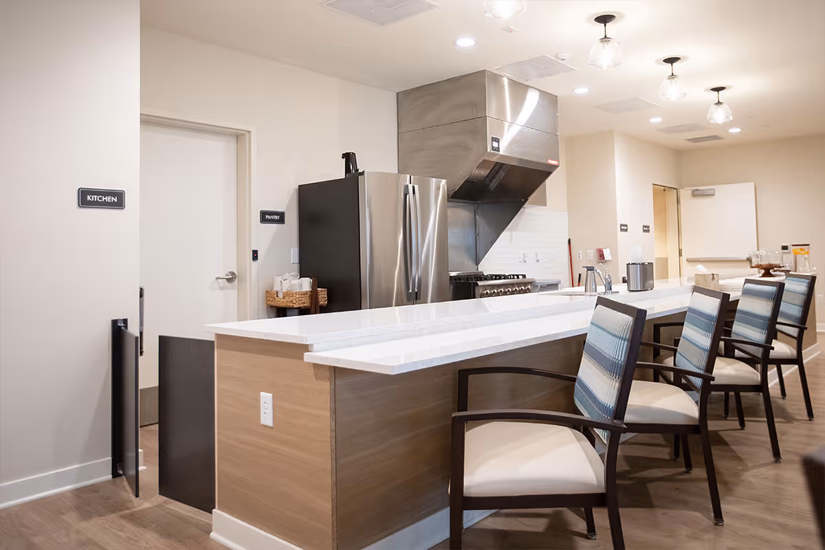 Bright communal kitchen with a long white island, bar chairs, stainless refrigerator and range hood.