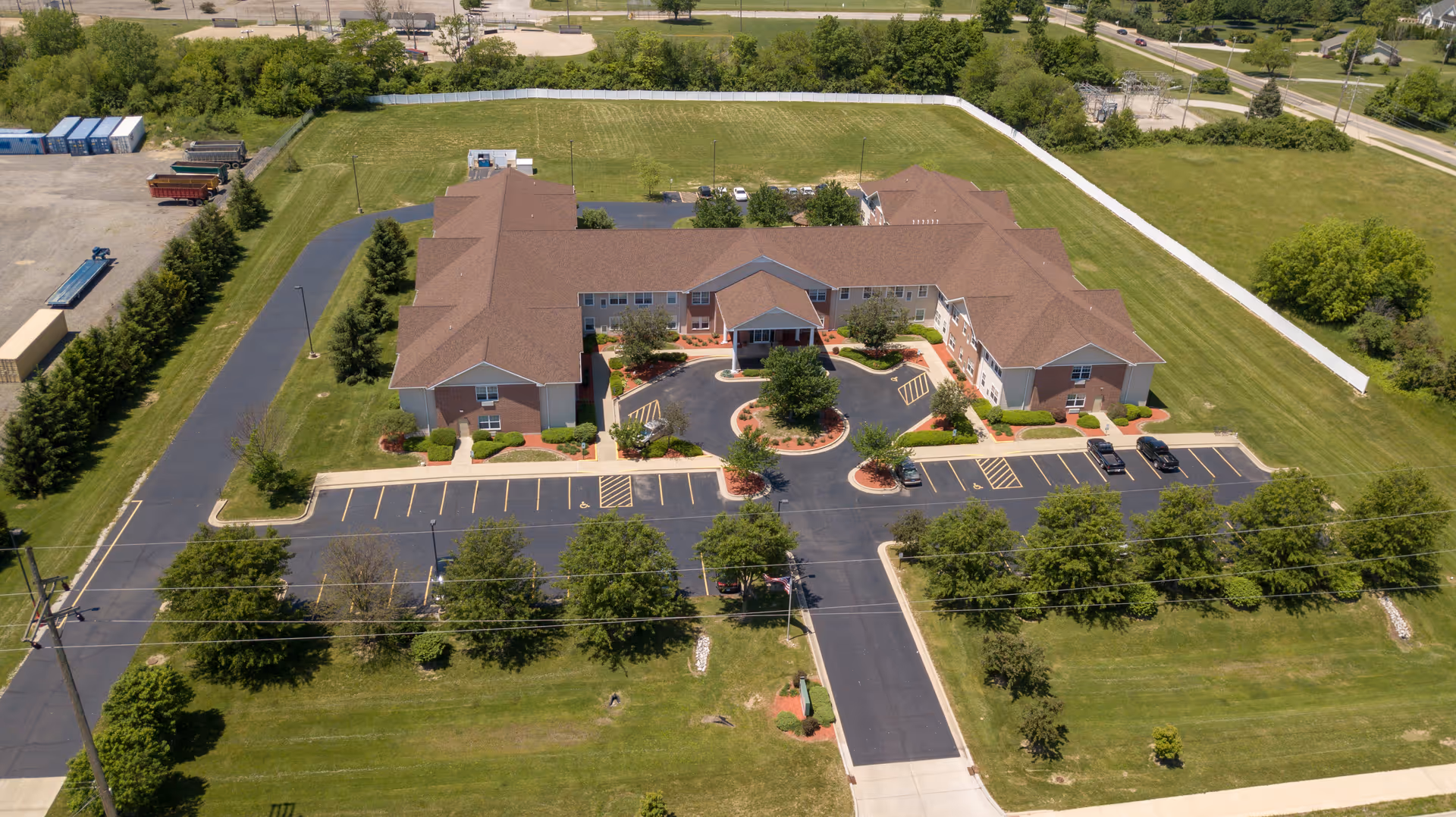 Aerial view of Bowman Estates of Danville, showing a large, single-story building with a brown roof surrounded by green lawns and trees. The building has a circular driveway with landscaped islands and parking spaces on both sides. The facility is bordered by a white fence and adjacent to a road.