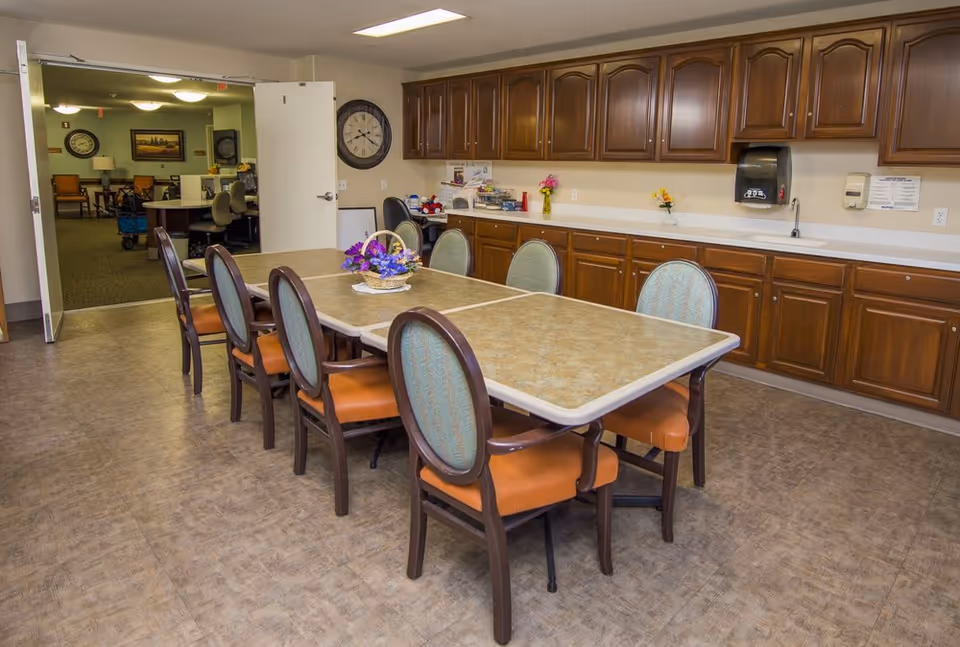 A room with a long rectangular table surrounded by eight chairs with orange seats and green backs. The table has a floral centerpiece. Along one wall, there are wooden cabinets with a countertop, a sink, a paper towel dispenser, and some small flower vases. In the background, an open double door leads to another room with more chairs, tables, and wall clocks.