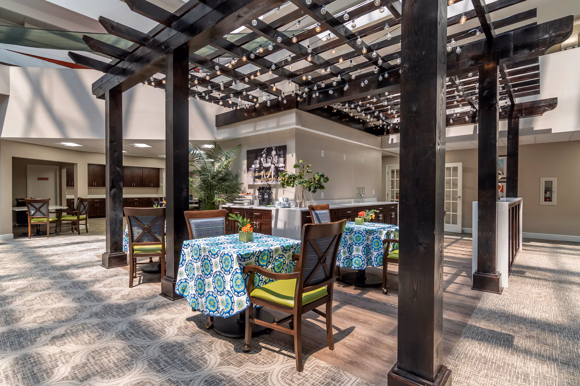 A bright indoor dining area with tables covered in blue and green patterned tablecloths, surrounded by wooden chairs with green cushions. The space features dark wooden beams overhead with string lights, a patterned carpet, and a counter with plants and coffee equipment in the background.