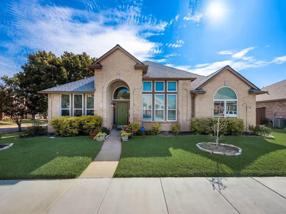Front exterior view of a single-story brick residential building with a green front door, large windows, manicured lawn, shrubs, and a clear blue sky with some clouds.