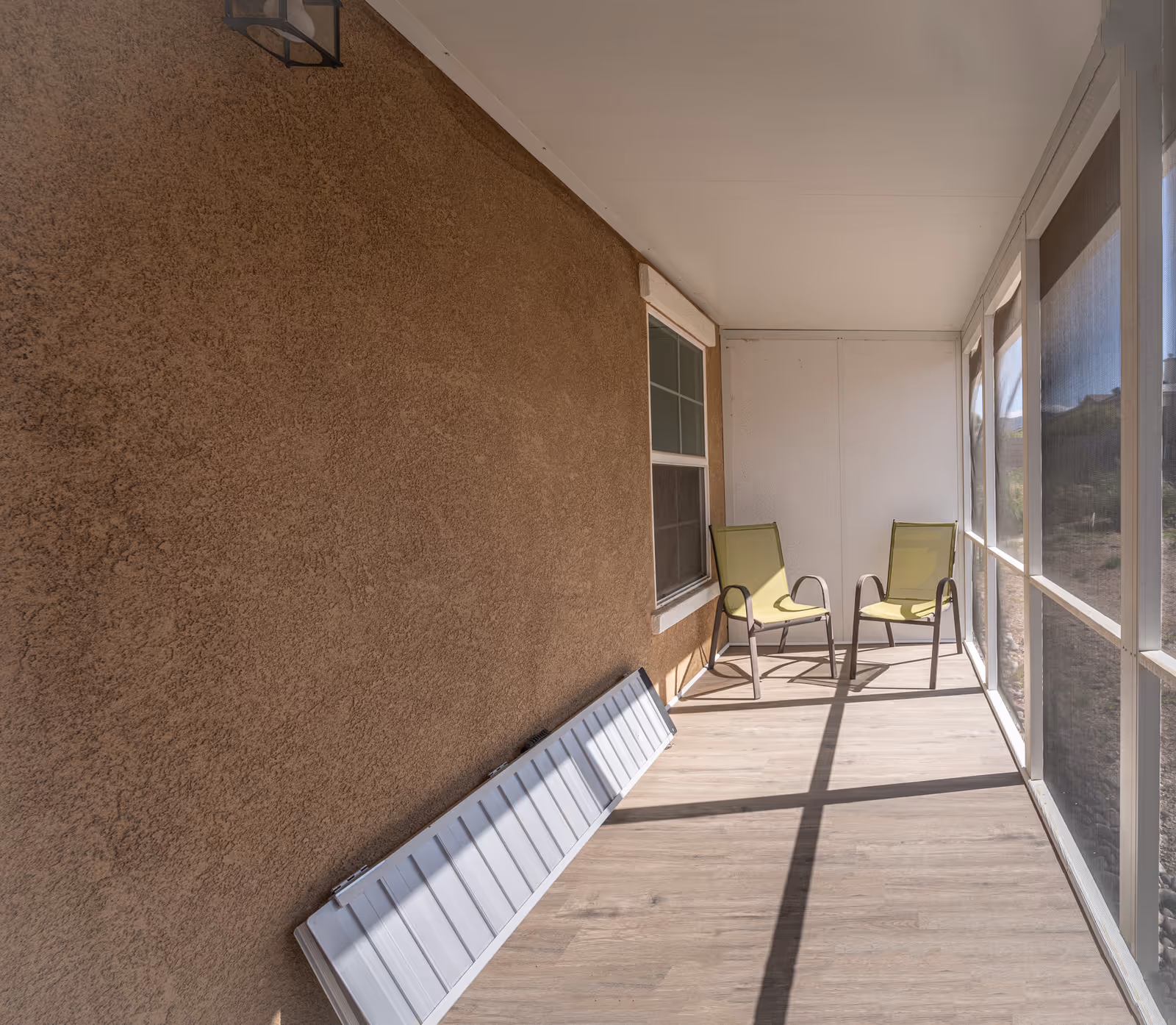 A screened-in porch with two green chairs, a stucco wall, and sunlight casting shadows on the floor.