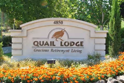 A curved stone entrance sign for Quail Lodge Retirement Community with orange flowers in front.
