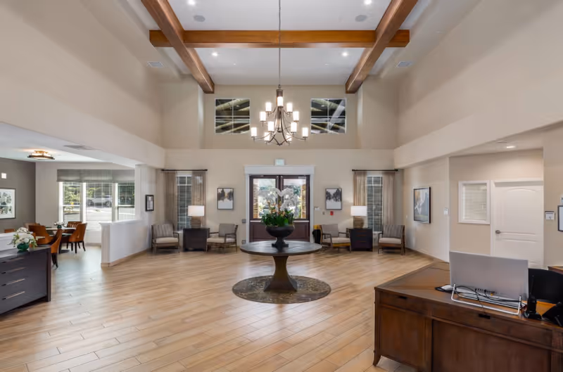 Spacious senior living lobby with high beamed ceilings, a central round table with flowers, chandelier, seating areas, and a reception desk.