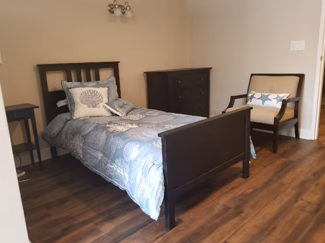 A small bedroom with a single dark wooden bed featuring blue and white seashell-themed bedding and pillows. Next to the bed is a small dark wooden nightstand, and against the wall is a matching dark wooden dresser. There is also a beige armchair with dark wooden arms and legs, decorated with a starfish-patterned pillow. The room has wooden flooring and light-colored walls.