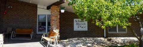 Brick building entrance with stone column, outdoor seating, sign, and leafy tree.