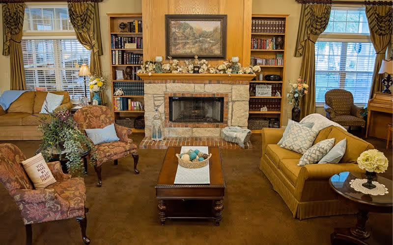 A cozy living room with a stone fireplace in the center, flanked by built-in wooden bookshelves filled with books and decorative items. The room features a mustard-yellow sofa with patterned pillows, two floral upholstered armchairs, a wooden coffee table with a decorative basket, and a side table with a flower vase. Large windows with gold curtains allow natural light to fill the space.