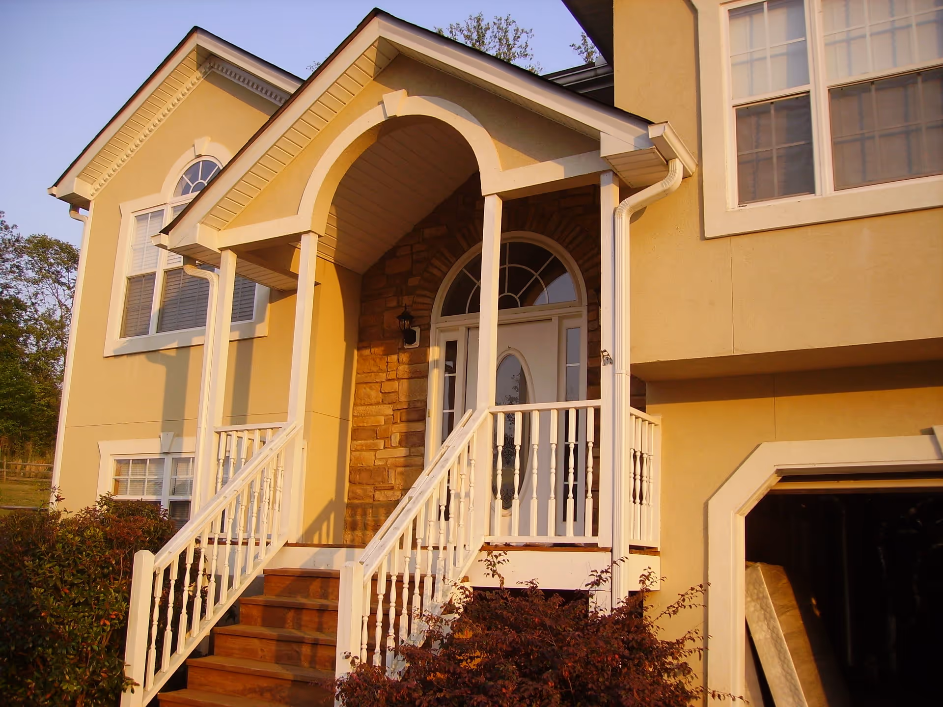 Front entrance of a yellow two-story house with white trim, a small porch with white railings, wooden steps leading up to a white door with an arched window above it, and surrounding bushes.