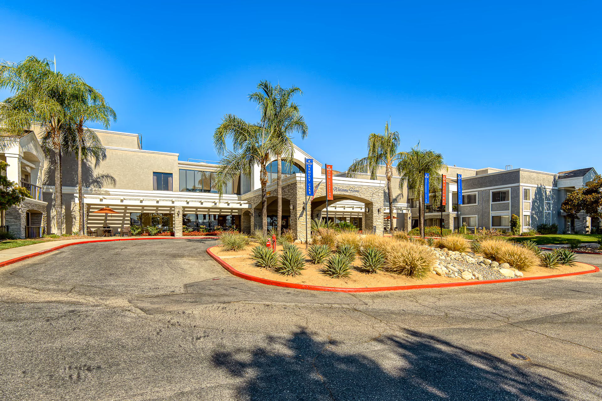 Front entrance and driveway of Menifee Senior Living with palm trees and a landscaped roundabout under a clear blue sky.