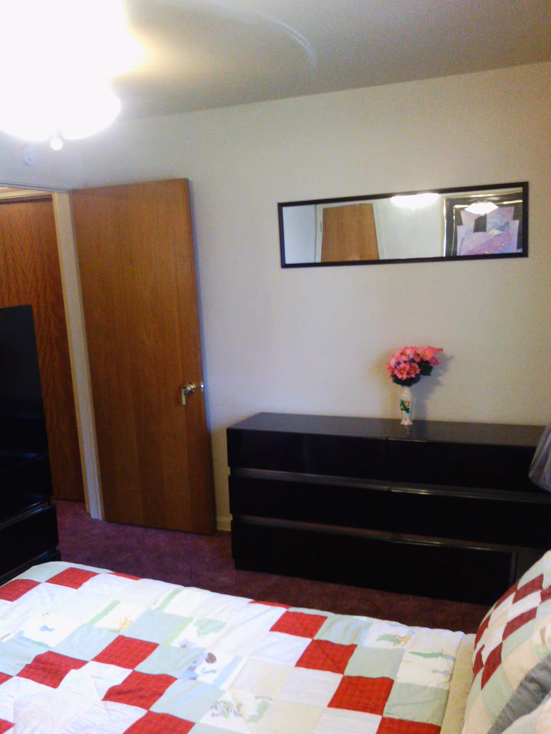 Bedroom with a quilted bed in the foreground, a black dresser topped with a vase of flowers, a wall mirror, and an open wooden door.