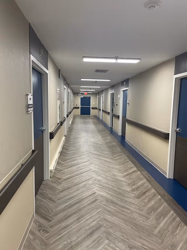 A long, well-lit hallway in a rehab center with multiple closed doors on both sides. The floor has a light wood pattern arranged in a herringbone style, and the walls are beige with dark handrails. The ceiling has several rectangular fluorescent lights and a smoke detector.