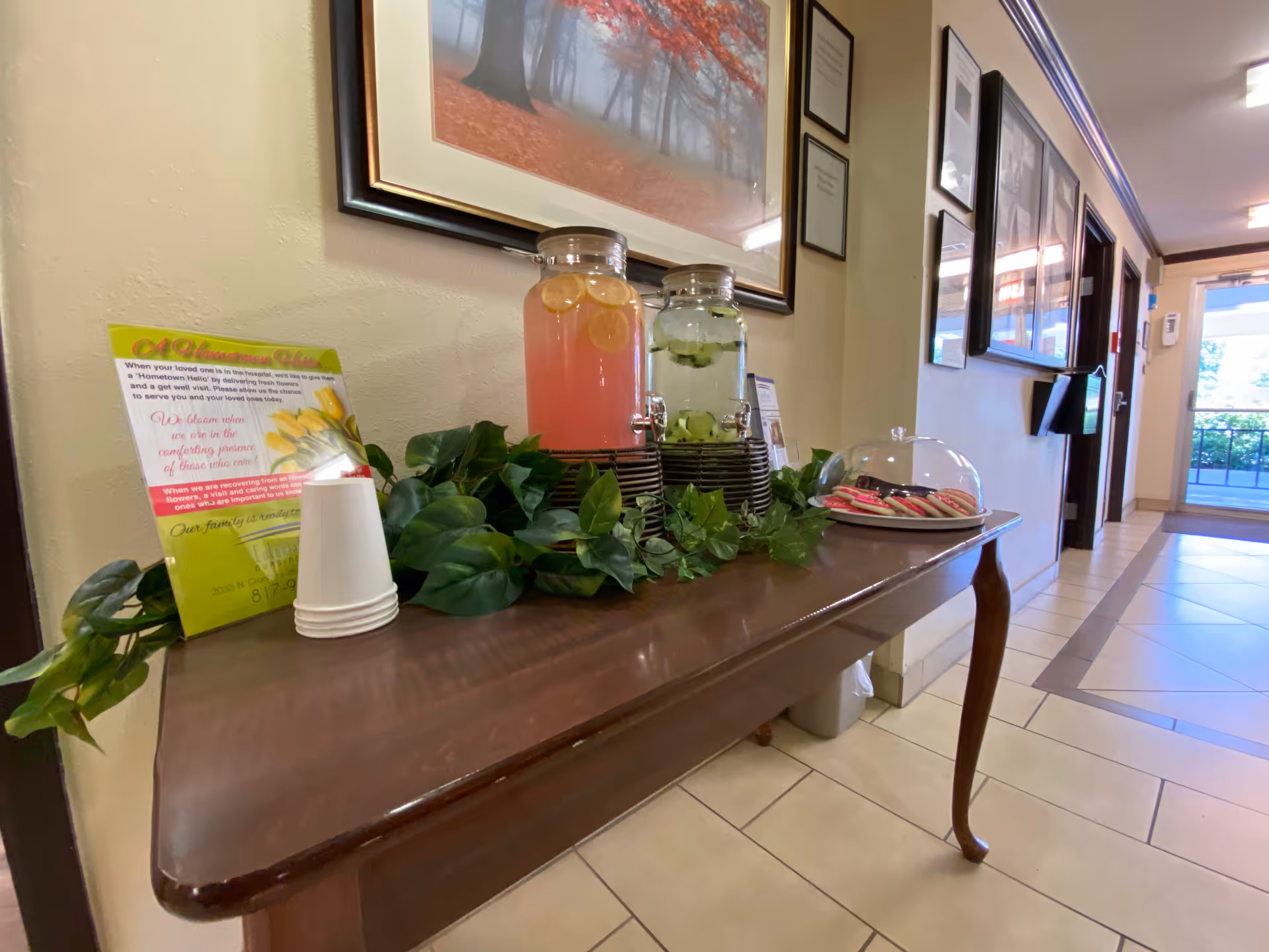 A wooden table in a hallway with two beverage dispensers, one with lemon water and the other with cucumber water, surrounded by green leafy decoration. There are stacked white disposable cups, a plate of cookies under a clear dome cover, and a framed picture of a tree with red leaves hanging on the wall above the table. The hallway has tiled floors and framed certificates or pictures on the walls, with a glass door leading outside at the end.