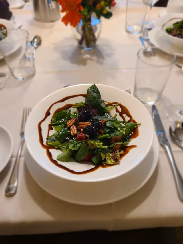 A white plate with a fresh salad consisting of mixed greens, blackberries, pecans, and a drizzle of balsamic glaze, placed on a white tablecloth. Surrounding the plate are clear drinking glasses, silverware, and a small vase with orange flowers in the background.