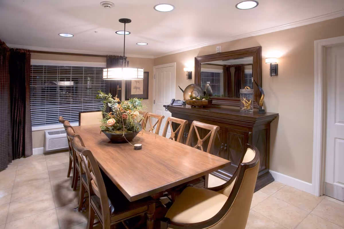 A well-lit dining room with a long wooden table surrounded by eight chairs. A floral centerpiece decorates the table. On the right side, there is a large wooden sideboard with decorative items and a large mirror above it. The room has beige walls, tiled flooring, and a window with blinds and dark curtains. Ceiling lights and wall sconces provide illumination.