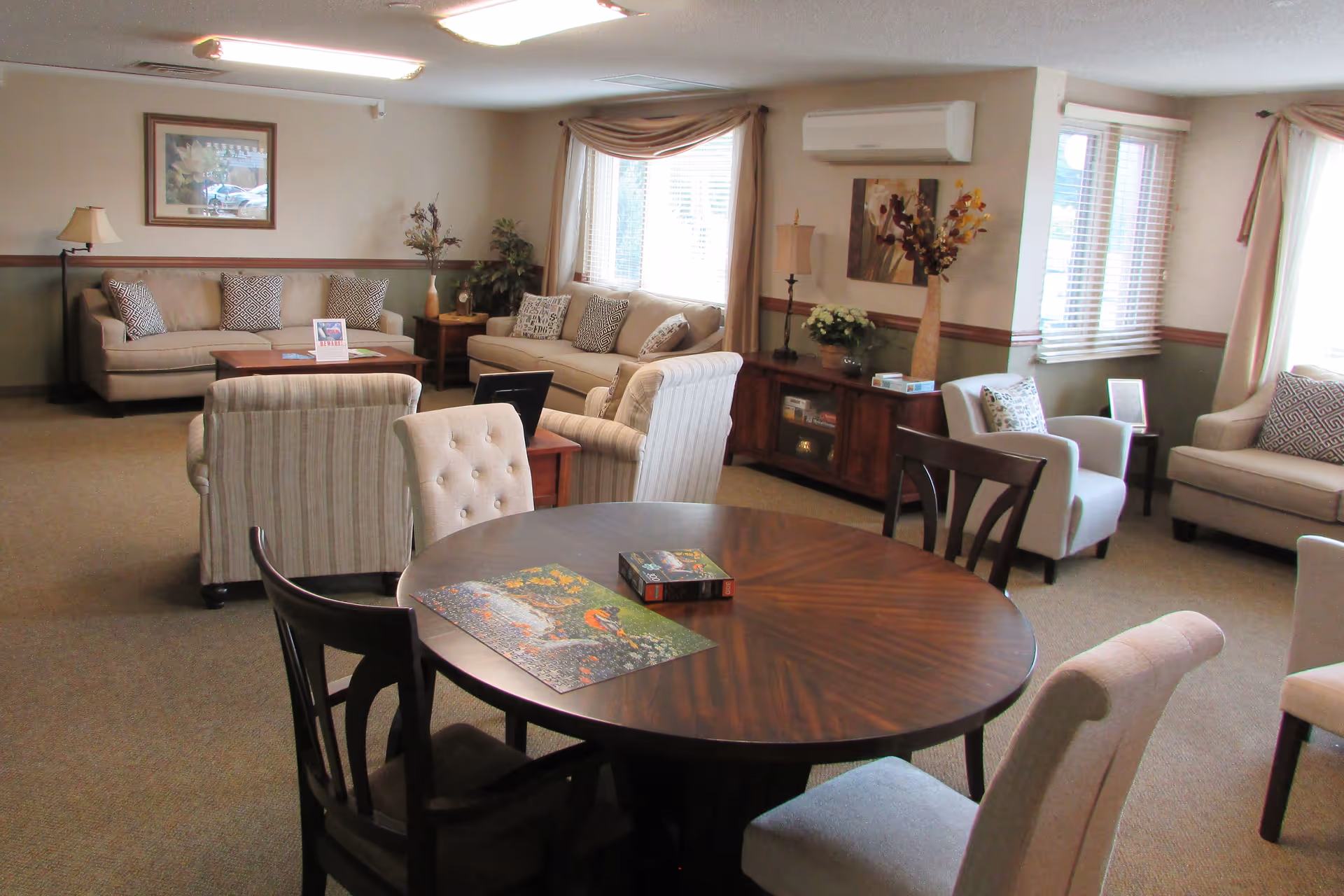 A cozy living room area with multiple sofas, armchairs, and a round wooden table with chairs around it. The room is decorated with framed artwork, lamps, plants, and a puzzle on the table. Large windows with curtains allow natural light to fill the space.
