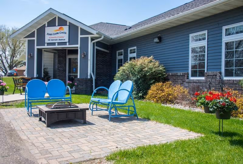 Outdoor patio area at Ashby Living Center featuring blue metal chairs and a fire pit on a paved surface, with the building entrance and landscaped bushes and flowers in the background.