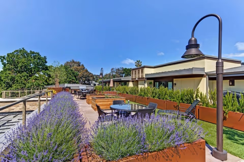 Outdoor patio area at Merrill Gardens at Lafayette featuring raised garden beds with lavender plants, a round glass table with chairs, lounge chairs, and modern street lamps under a clear blue sky.