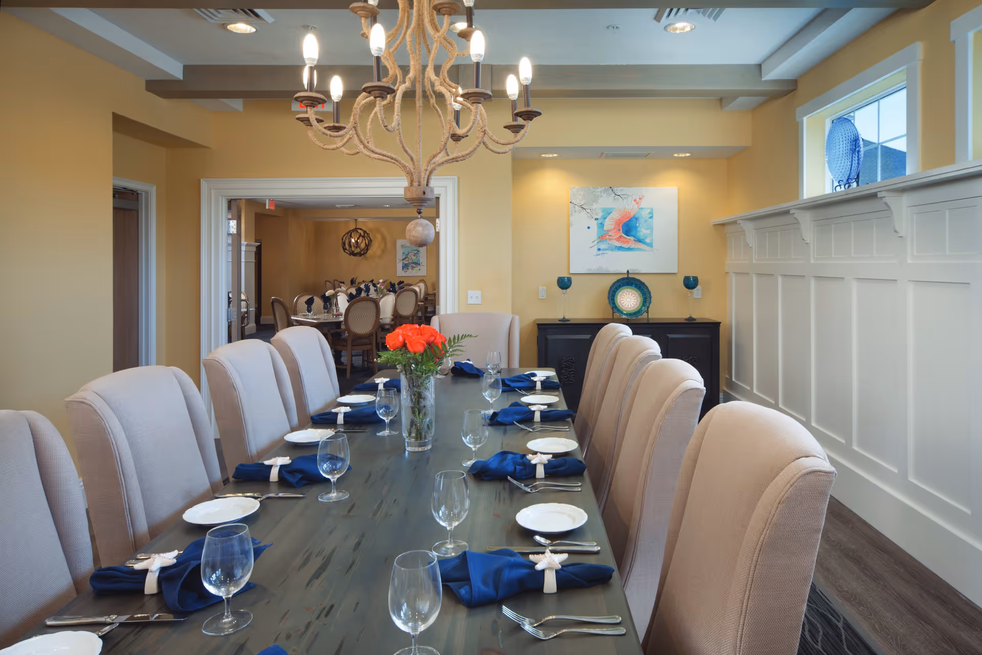 A formal dining room with a long wooden table set for a meal. The table is arranged with white plates, blue napkins with starfish napkin rings, silverware, and empty wine glasses. A vase with red flowers is placed in the center of the table. Beige upholstered chairs surround the table. The room has yellow walls, white wainscoting, and a large window with a blue decorative plate on the ledge. A rope chandelier hangs above the table. In the background, another dining area with similar chairs and tables is visible through a wide doorway.