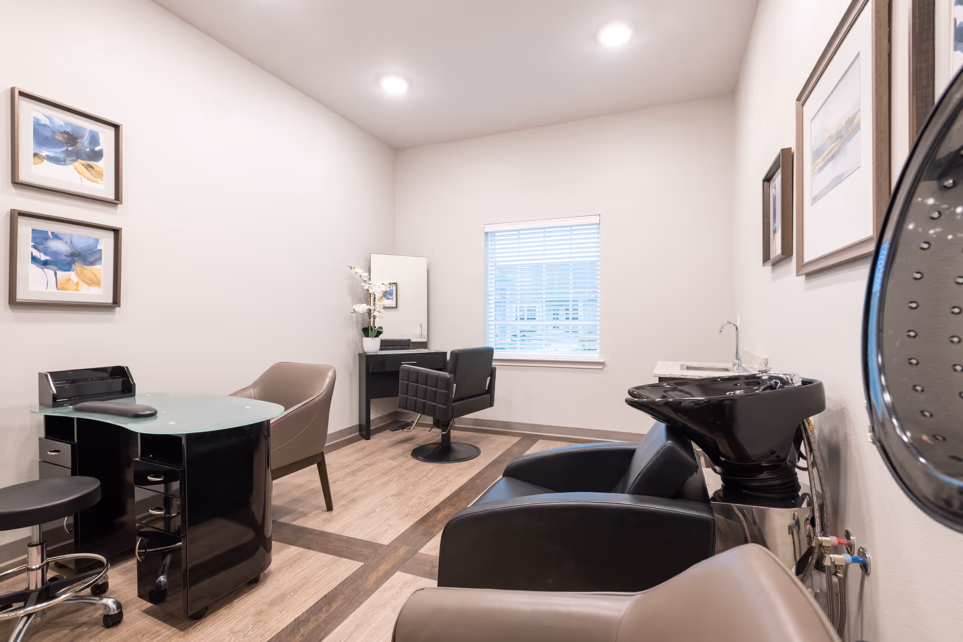 Interior of a salon room with a black hair washing station, a styling chair in front of a mirror, a manicure table with chairs, and framed artwork on the walls. The room has light-colored walls and wood flooring with a window letting in natural light.