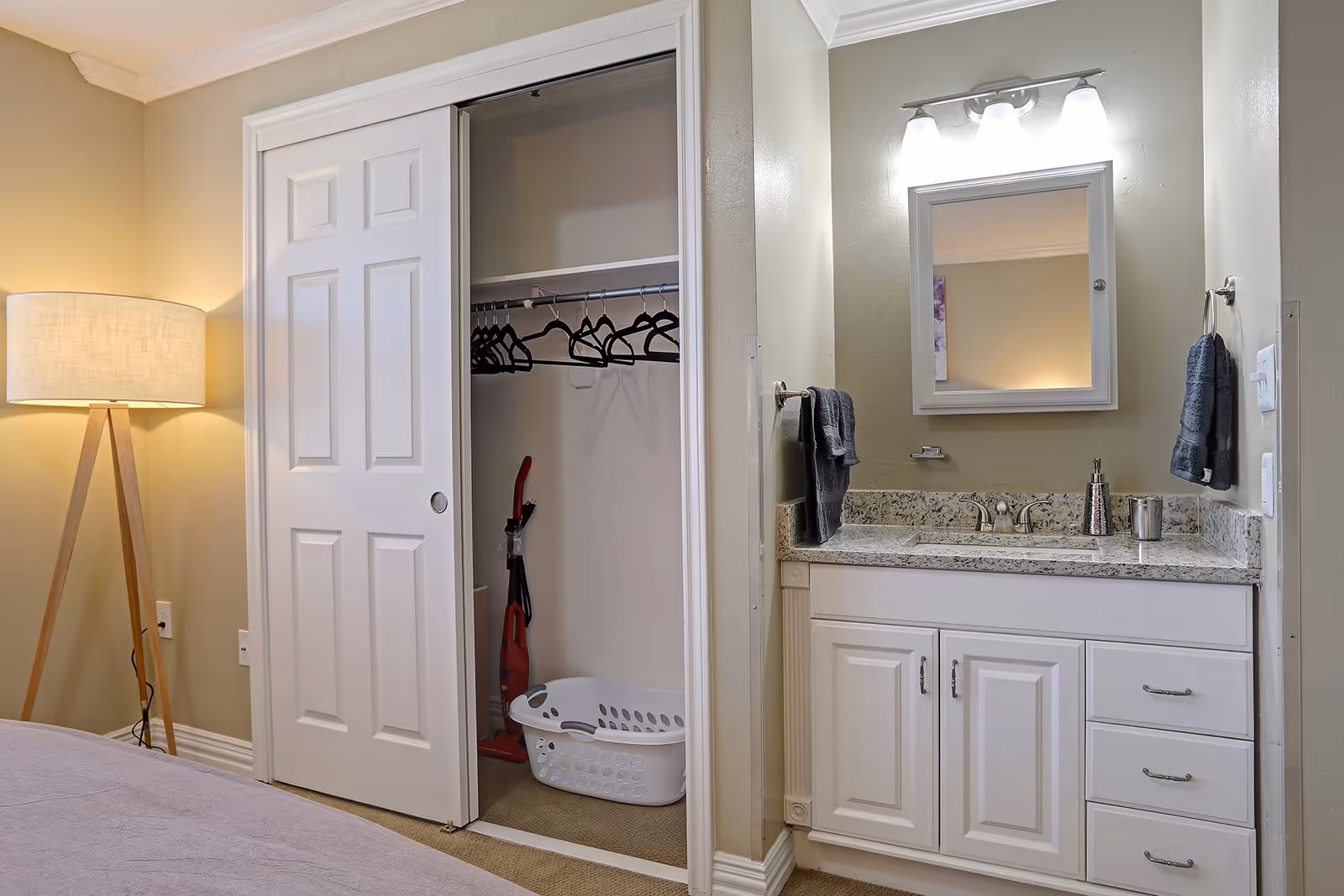 Interior view of a room featuring a white sliding closet door partially open revealing a closet with hangers, a vacuum cleaner, and a laundry basket. To the right is a bathroom vanity with a granite countertop, a sink, a mirror with lights above it, and two dark gray hand towels hanging on either side. A tripod floor lamp with a white lampshade is positioned to the left of the closet.