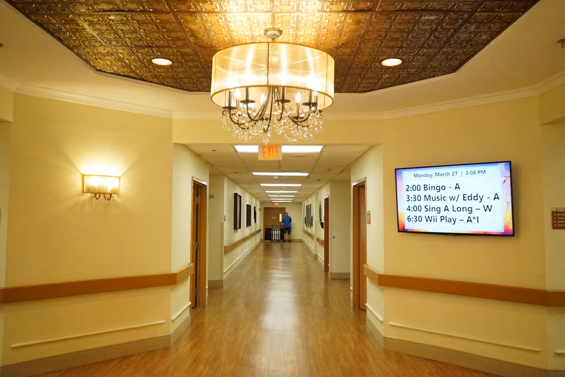 Well-lit interior hallway of a senior living facility with wood floors, wall handrails, a decorative ceiling light, and a mounted TV showing activities.