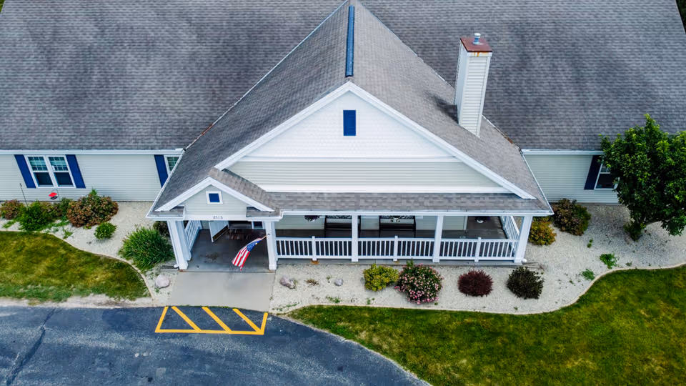 Aerial view of the front entrance of a single-story senior living facility building with a covered porch, white railings, and an American flag. The building has a gray roof, white siding, blue window shutters, and landscaped bushes and grass surrounding the entrance.