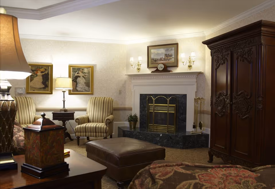 A cozy living room with a fireplace featuring a brass screen and a marble hearth. The room has two striped armchairs, a large dark wooden armoire with intricate carvings, a brown leather ottoman, and a wooden side table with a decorative lamp. The walls are adorned with framed paintings and wall sconces providing warm lighting.