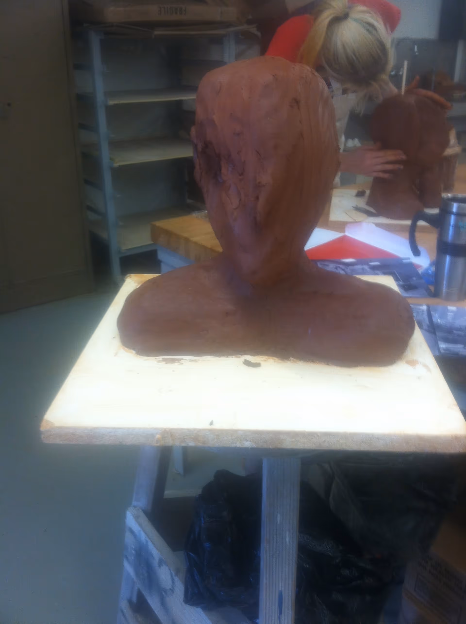 A clay sculpture of a human head and shoulders on a wooden board in an art studio. In the background, a person is working on another clay sculpture, and shelves and art supplies are visible.