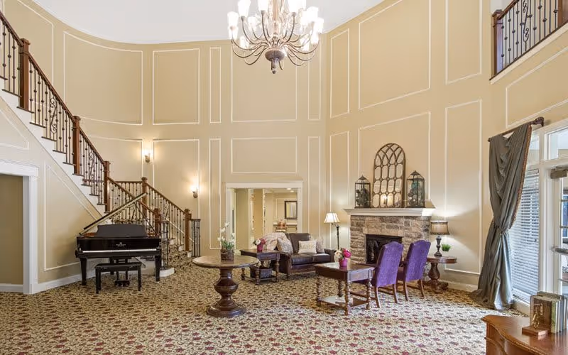 A spacious and elegant living room area with high ceilings and beige walls featuring white trim. The room includes a grand piano near a staircase with wooden railings, a round wooden table with a plant, a seating area with a sofa and purple chairs around a small table, and a stone fireplace decorated with lanterns and a mirror. Large windows with curtains allow natural light to enter the room.