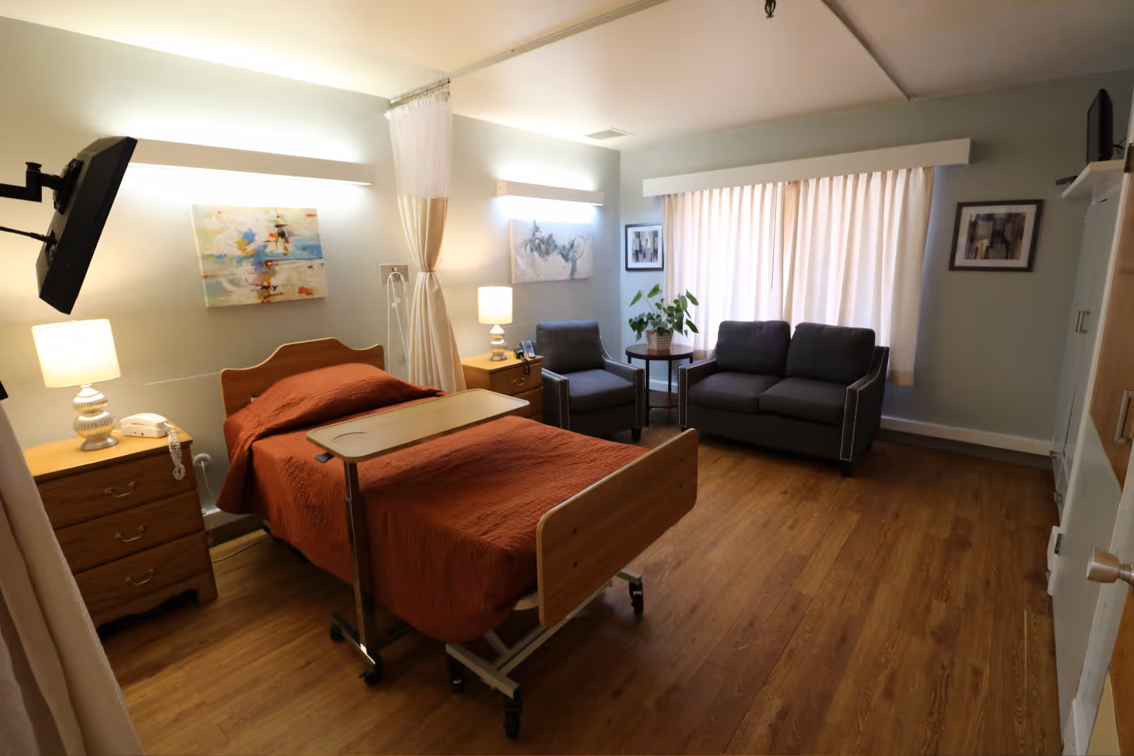 A furnished patient bedroom with a hospital bed and overbed table, nightstands and lamps, and a seating area with two chairs and a sofa.