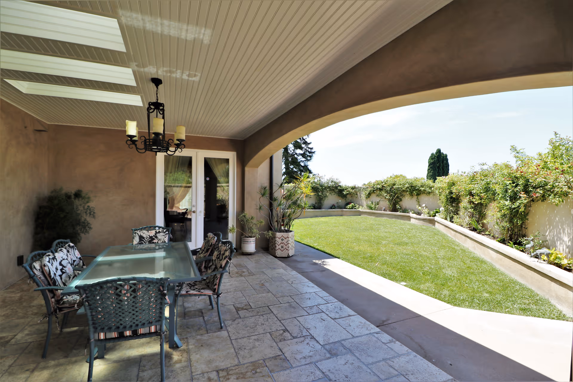 Covered outdoor patio area with a glass-top dining table and six cushioned chairs. The patio has a tiled floor, a chandelier hanging from the ceiling, and potted plants near a set of double glass doors. Beyond the patio is a well-maintained grassy yard bordered by a low wall with climbing plants and shrubs.