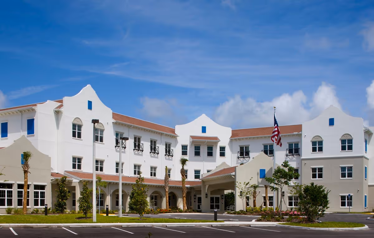 White three-story Mediterranean-style senior living building with an arched entrance, landscaped front, and an American flag under a blue sky.