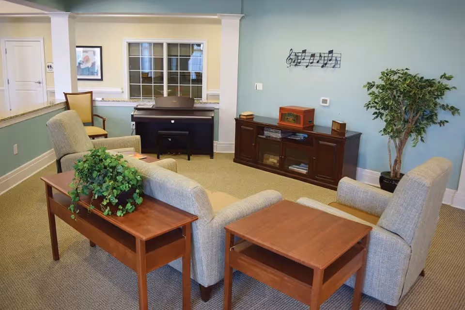 A cozy living room area with two beige armchairs and a matching loveseat arranged around two wooden side tables. A wooden console with a vintage-style radio and books sits against a light blue wall decorated with a musical notes wall art. A potted plant is placed in the corner, and a piano with a chair is positioned near a window with white trim. The room has beige carpeting and light-colored walls.