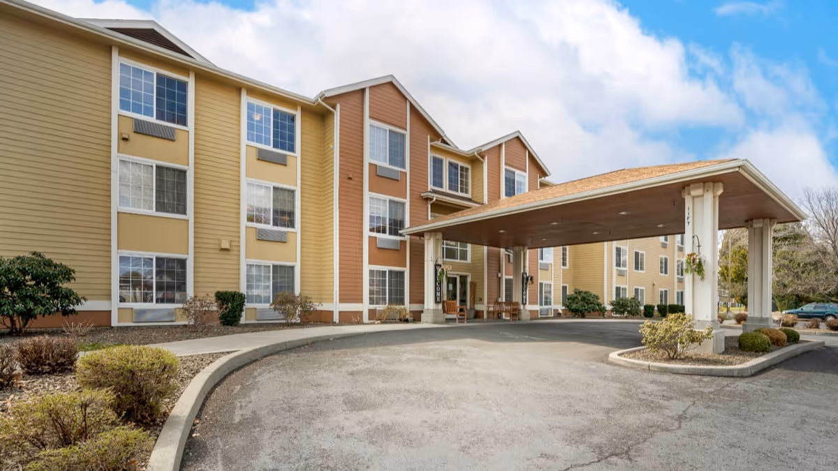 Exterior view of a three-story senior living facility building with beige and brown siding, multiple windows, and a covered entrance with columns. There are shrubs and small bushes along the driveway leading to the entrance under a partly cloudy sky.