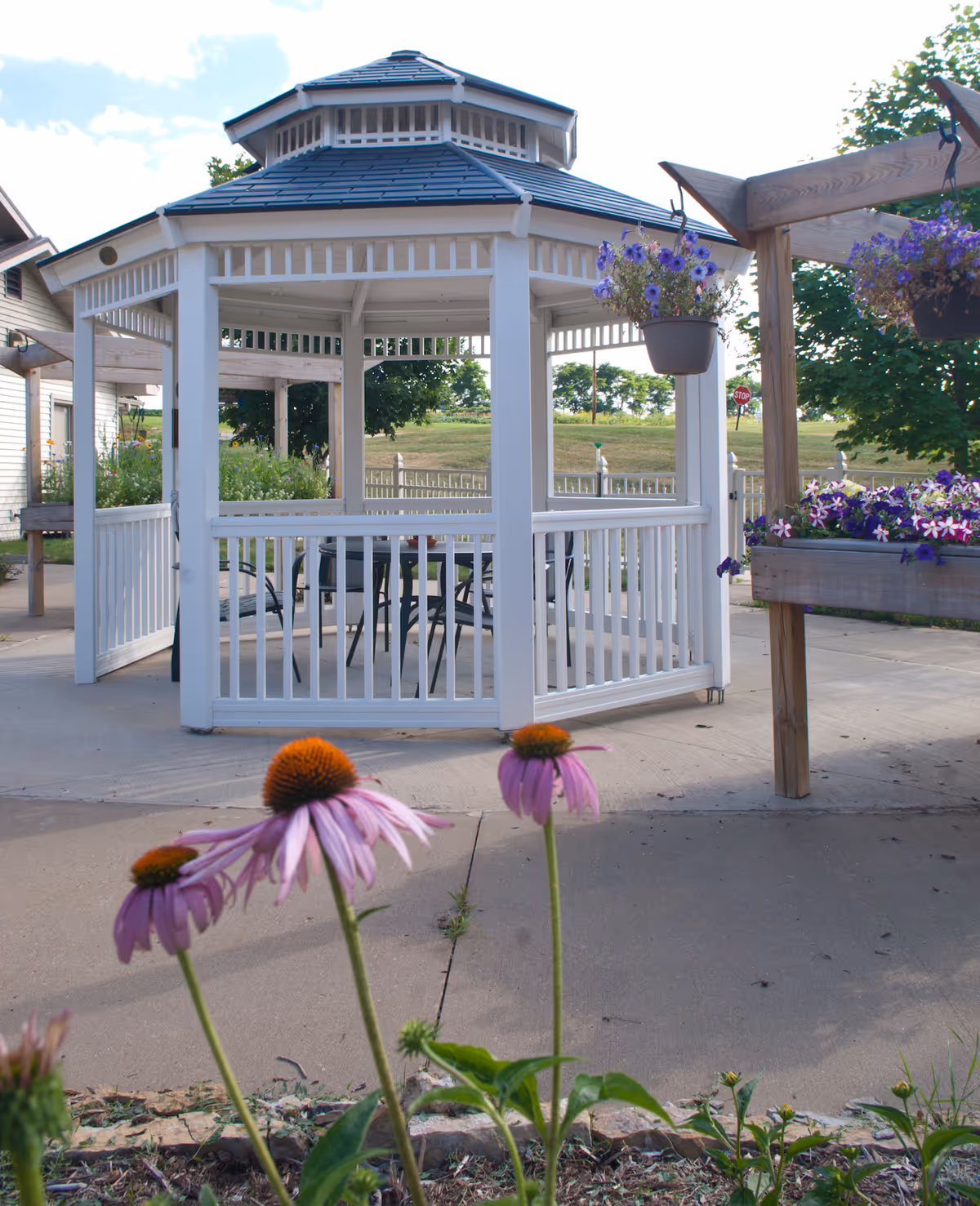 White octagonal gazebo with outdoor chairs, hanging flower baskets, and purple coneflowers in the foreground.