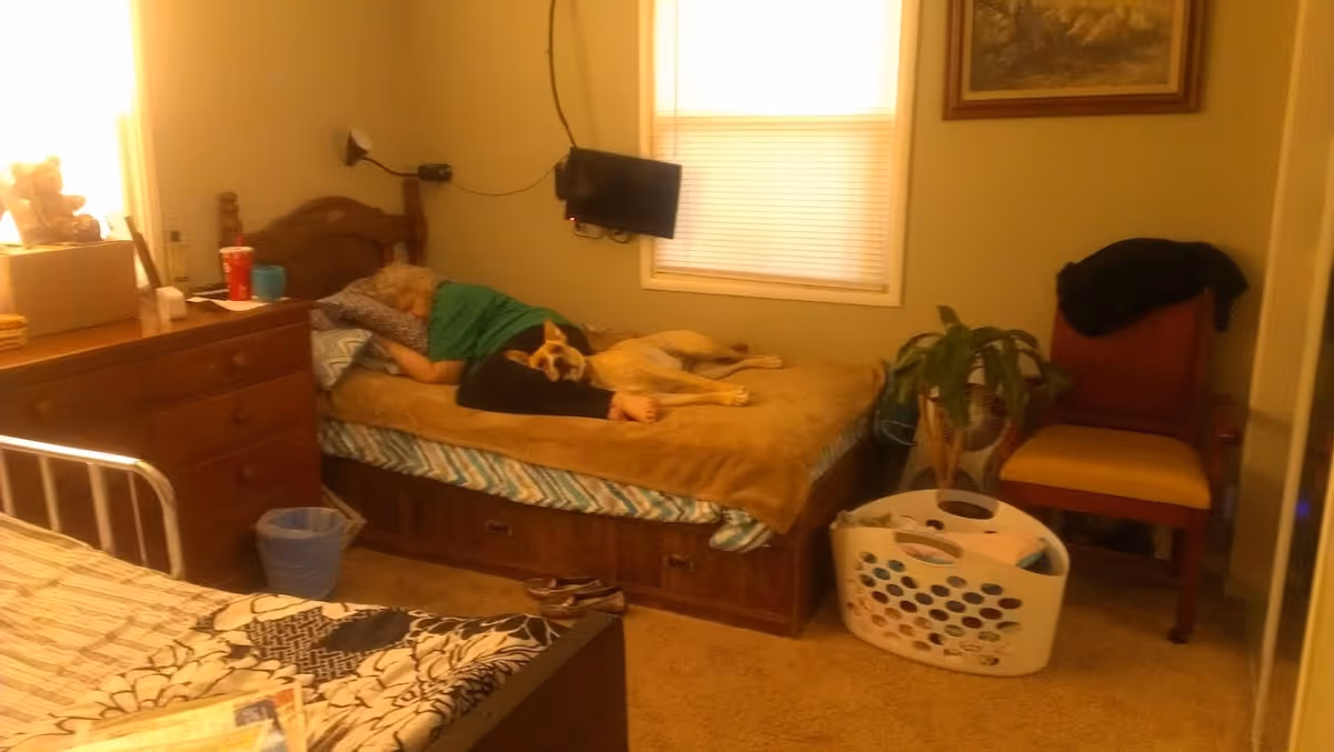 Person lying on a bed with a dog in a furnished bedroom containing a dresser, chair, and laundry basket.